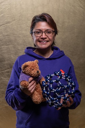 A picture of Dr Pooja Goddard holding a teddy bear and a model of a 'buckyball' (a type of catalyst)