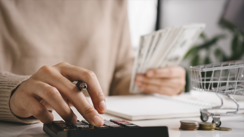 A close-up photograph of a person holding cash notes in one hand while using a calculator with the other hand.