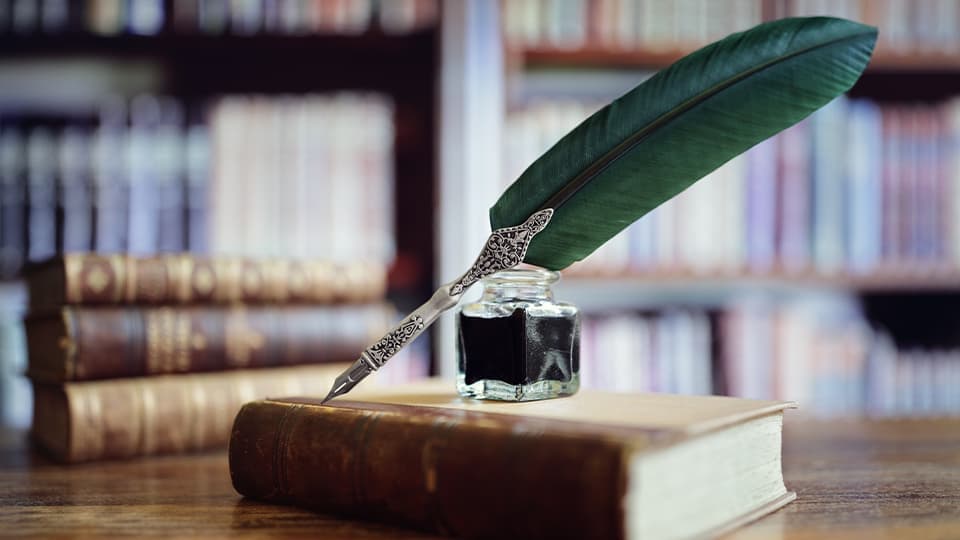 A green quill pen rests in a glass inkwell on top of an old hardcover book, set against a blurred background of library bookshelves.
