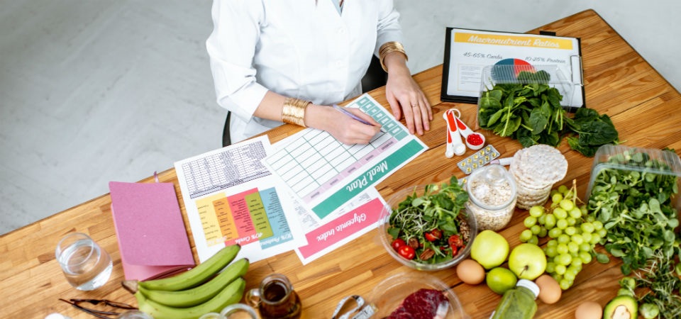 a woman sitting at a table writing on meal planner sheets, there is a selection of vegetables and fruit spread out
