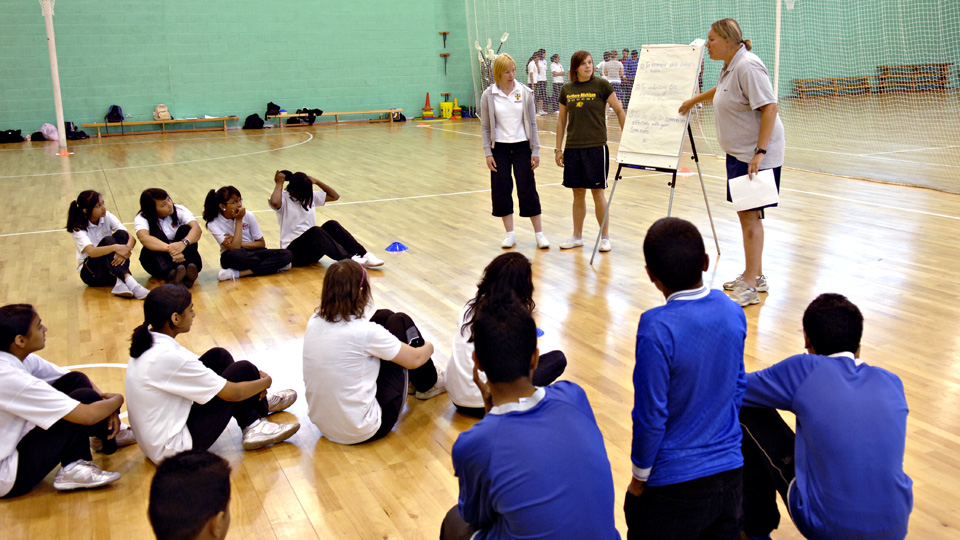 a group of pupils sitting on the floor listening to the 3 PE teachers