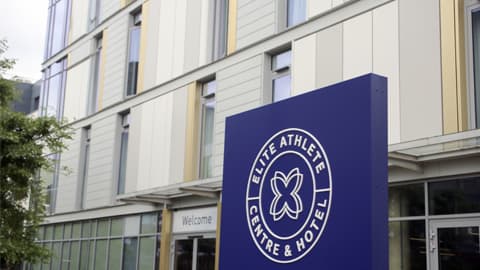 Modern exterior of the Elite Athlete Centre & Hotel featuring a tall blue sign with the hotel’s circular logo and name.