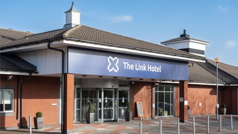 Front view of The Link Hotel featuring a wide blue sign with the hotel name and logo above glass double doors, set against red‑brick walls with a tiled roof and clear blue sky in the background.