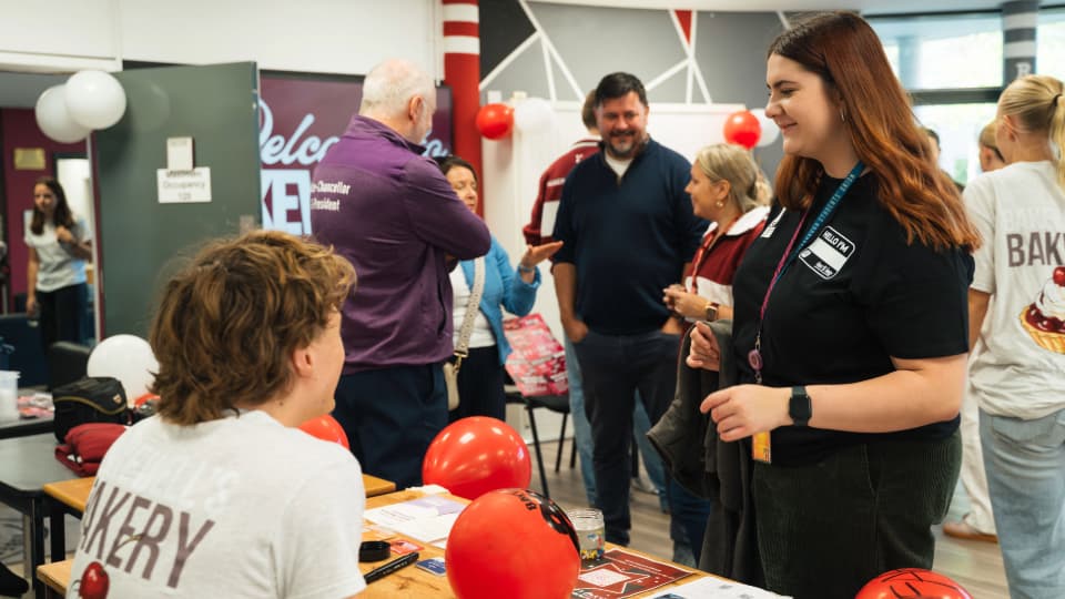 People speaking with visitors at a university information event. Tables decorated with red balloons and promotional materials are set up in an indoor venue, with attendees chatting and collecting information from student representatives.