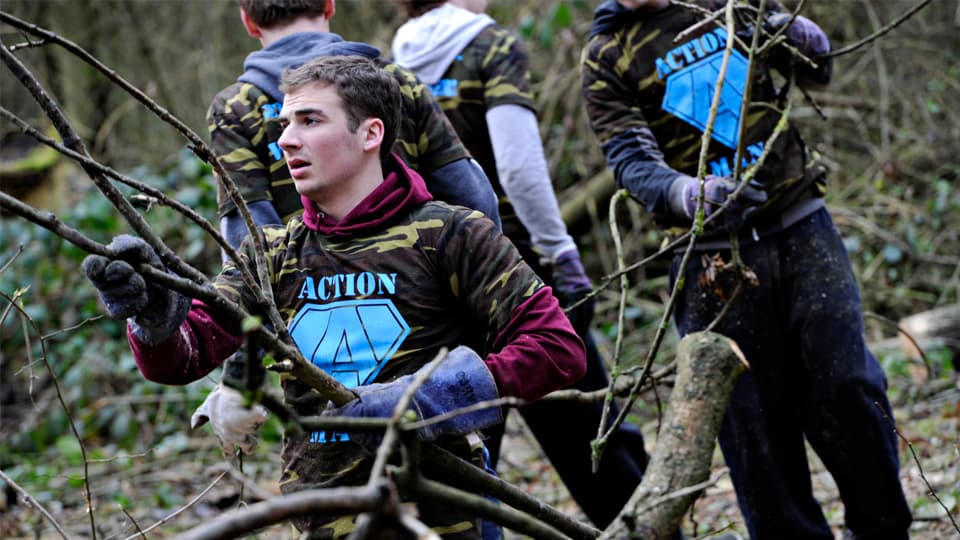 Several volunteers wearing camouflage-patterned shirts with a blue “ACTION” logo clear branches and fallen wood in a wooded area. One volunteer in the foreground carries a large branch while others work among trees and undergrowth in the background.