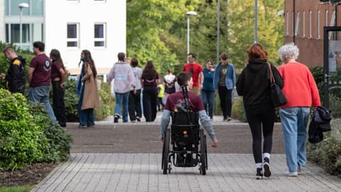 A group of students and visitors walking along a paved pathway between university buildings. In the foreground, a student in a wheelchair is moving forward, accompanied by two people walking beside them. Other students are walking and talking in the background, with greenery and modern campus buildings on either side