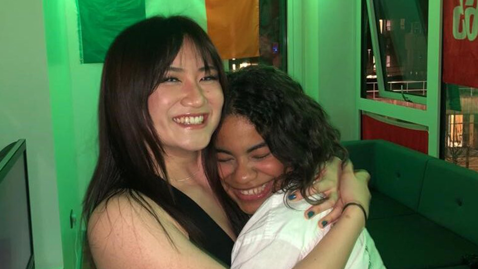 two female students hugging and smiling in a bedroom in cayley hall, there are posters on the wall and the room is lit by a green led tinted light.