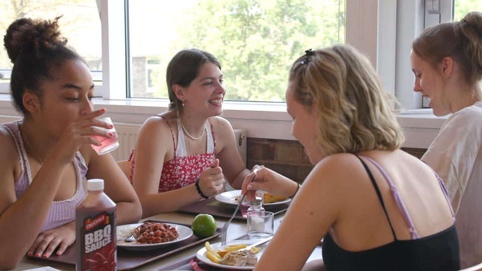 four students sat at dining table in dining hall eating fresh nutritious student meals and laughing together