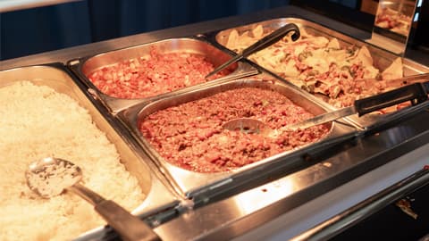 Close-up of a hot food counter featuring stainless steel trays filled with cooked dishes. The trays contain white rice, a minced meat chili dish, diced vegetables, and nachos topped with melted cheese and garnishes. Serving spoons and tongs rest in the trays.