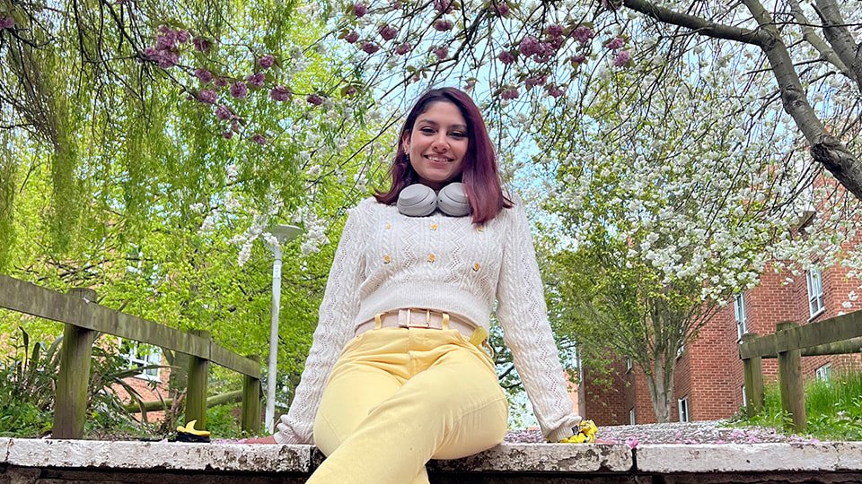 a female student sitting outside on a spring day smiling toward the camera, there are many trees surrounding her with white and purple blossom.