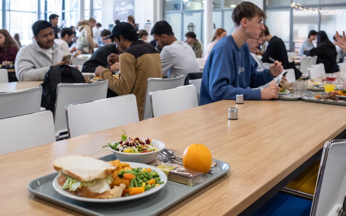 A cafeteria setting with a long wooden table and trays of food. The nearest tray includes a large sandwich on white bread, mixed vegetables, a colorful salad with dressing, a whole orange, and a wrapped brownie. Other trays with meals and drinks are visible on surrounding tables, and groups of people are seated and eating in the background.