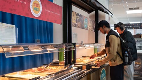 Two people serving themselves food from a self-service counter in a cafeteria. The counter has glass shields and trays filled with hot dishes, including rice and pasta options. Overhead signs display ‘Tex Mex’ and ‘The Pasta Bar,’ and the counter has wooden panels with matching signage.”