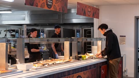 Cafeteria food counter with trays of hot dishes under a glass shield. A person is serving food onto a plate, while staff members stand behind the counter in a commercial kitchen setting. Overhead signs display the theme ‘Wholesome & Hearty,’ decorated with food illustrations.