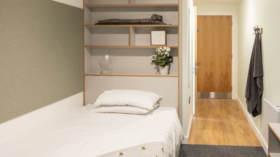 Modern student bedroom with a single bed, built-in shelving, wood flooring, and a doorway leading to an en-suite area. Decorative items and a potted plant are displayed on the shelves above the bed.