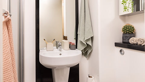 Stylish modern private en-suite bathroom in bedrooms at Stratford One accommodation. There is a shower on the left, a sink with toothbrushes, soap and a large mirror in the centre of the room, and a toilet with toilet paper on the right.