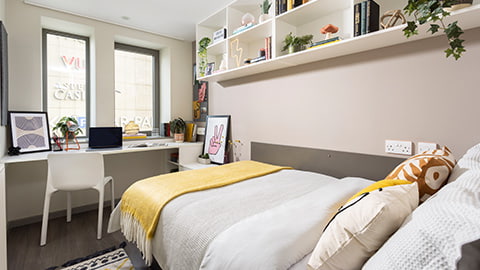 Classic en-suite bedroom at Stratford One, student accommodation at Loughborough London campus. The bed is a small double with cosy bed covers, duvet, and yellow blanket. There is a spacious desk, with seating, shelves, bedside table and plenty of storage space. The room is modern and ideal for students with incredible city views.