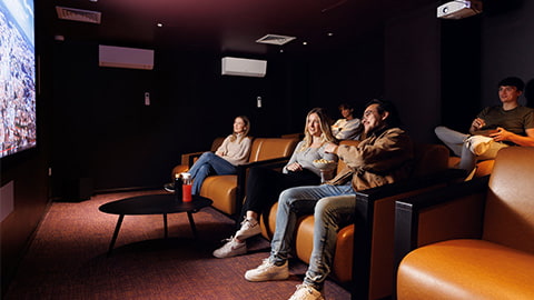 Comfy cinema room inside Stratford One Loughborough London accommodation. There is a group of students sitting on large comfy sofas all sat facing a large cinema screen with dim room lighting.
