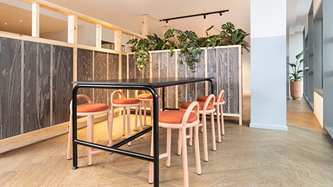 Modern, light and spacious social and study space in Stratford One Loughborough London student accommodation. There is a spacious long study table with rows of comfy stalls sat alongside the study table. The study space is sectioned off from other social spaces by walls of plants.