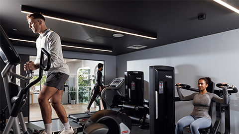 Fitness studio in Stratford One Loughborough London accommodation. There is a range of sporting and fitness equipment in the fitness studio gym, including a student running on a treadmill, a student on an elliptical cross-fit trainer, and a student using a weight training machine.