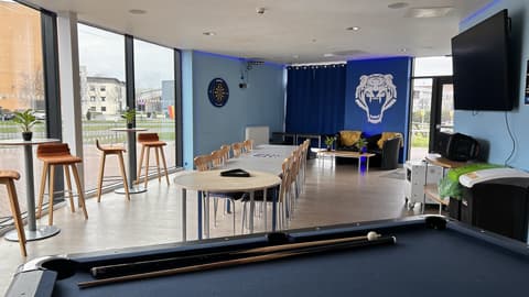 Modern student common room with pool table, large windows, bar stools, study tables, sofas, and a blue feature wall with a tiger graphic.