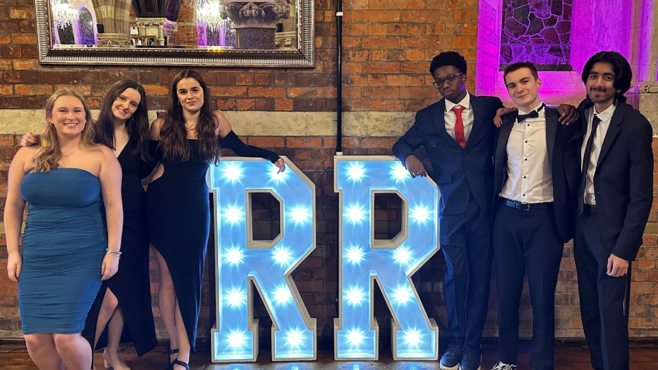 Alt text: A group of six students dressed formally stand beside illuminated “RR” letters at a social event, posing and smiling in front of an exposed brick wall with purple accent lighting.