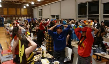 Students wearing a variety of costumes sit and stand around long tables in a large indoor hall, taking part in an organised group activity.