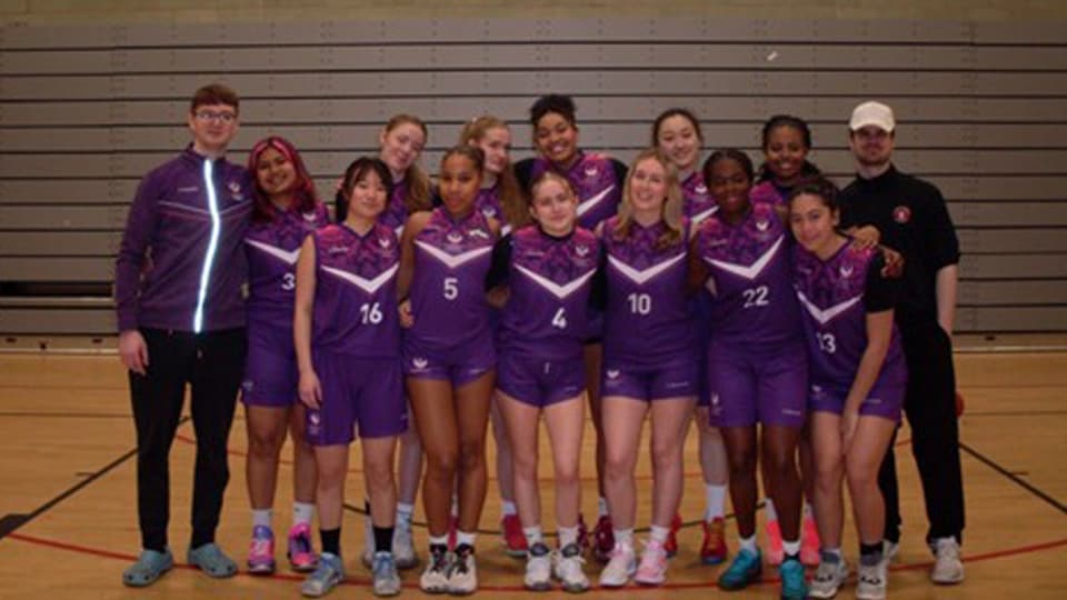 Group of indoor basketball players and two coaches standing together on a wooden sports hall court, wearing matching purple kits with white detailing, posing in front of a lined wall backdrop.