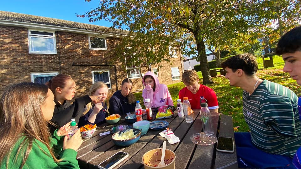 A group of students sitting around an outdoor picnic table sharing food and drinks on a sunny day, with brick accommodation buildings and a tree with autumn leaves in the background.
