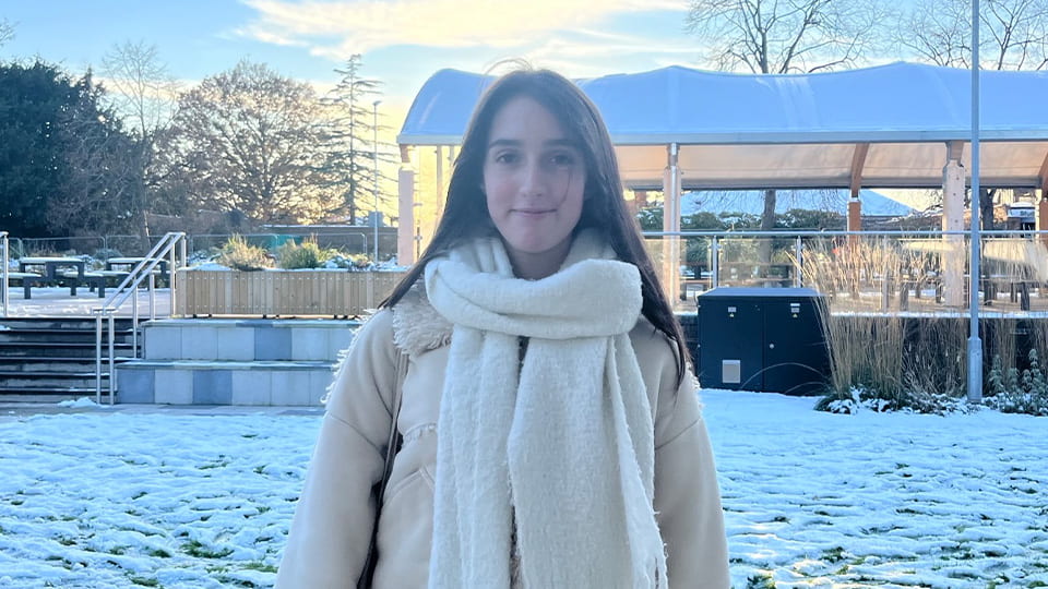 Student facing camera and smiling, she is wrapped up warm in winter coat and scarf. Behind her campus is covered in snow.