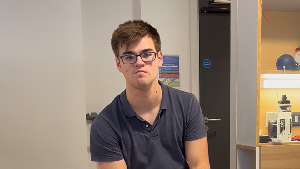 Student leans forward in wheelchair to give a light smile to the camera. He is sat in his bedroom at Claudia Parsons.