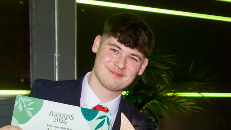 Loughborough University student smiles towards the camera, he wears a suit and is holding an award for his hard work on the Butler Court hall committee.