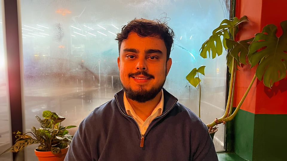 Student smiling at camera sat in a cafe with his back against steamed glass wall and indoor plants
