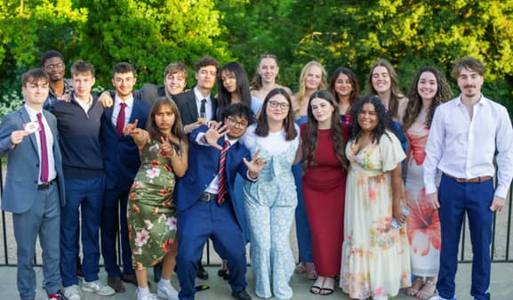 A large group of university students in formal outfits pose together outdoors, smiling and celebrating a summer ball.