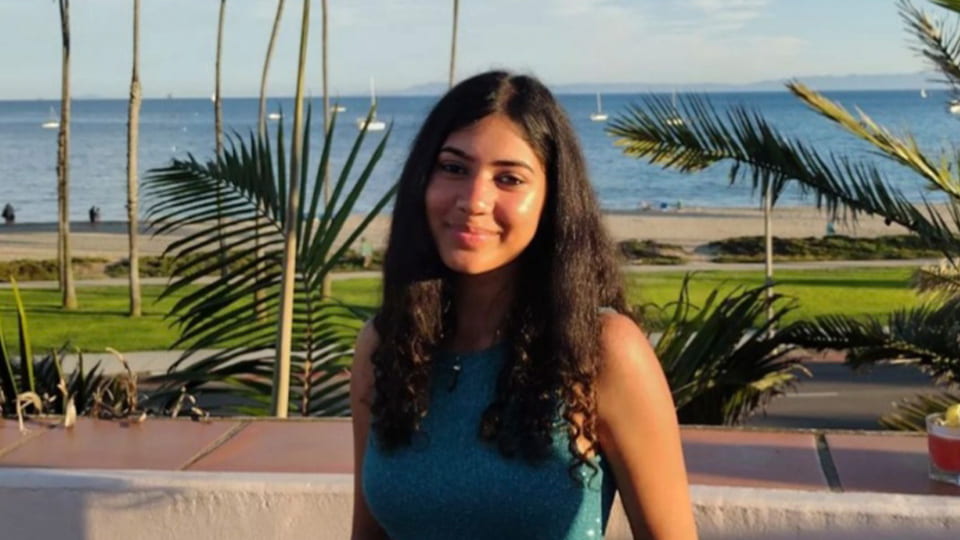 Female student turns to face the camera and smiles, she is standing outside at a holiday resort and the sun shines down on her.