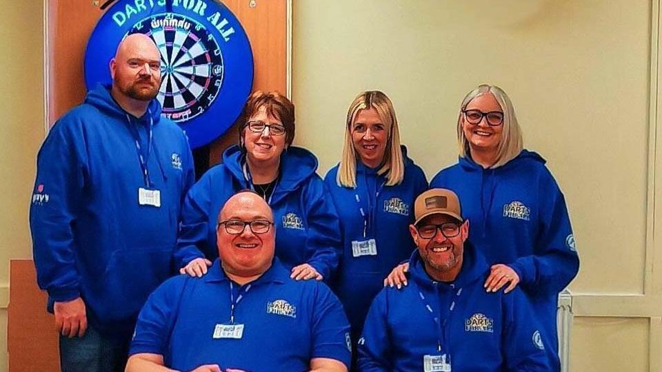 Six adults wearing matching blue hoodies and ID badges pose together in front of a dartboard with a 