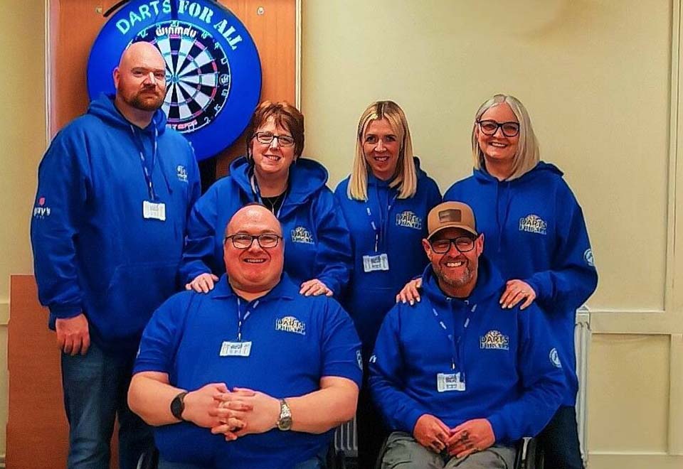 Six adults wearing matching blue hoodies and ID badges pose together in front of a dartboard with a 