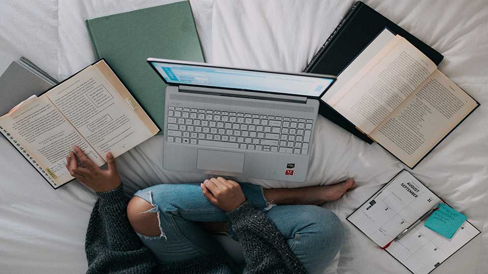 A student sitting on a bed, with a laptop, books and notebook around her