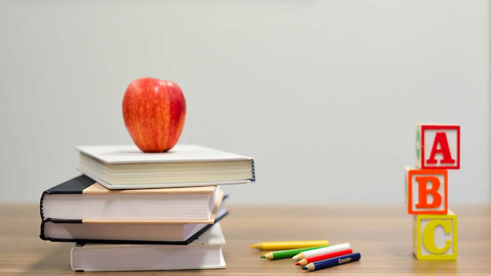 An apple on a few books, pencils next to it and A, B, C building blocks next to that. All of them on a table against a white background