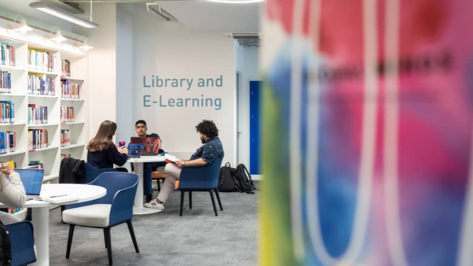 Three students around a round table with the words 'library and e-learning' on the wall