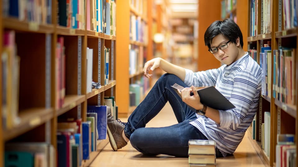 Young man with short dark hair and glasses reading a book whilst sat between two shelving units filled with books