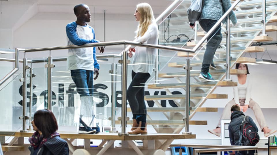 Two students chatting on staircase of level 3 in Pilkington Library, featuring self service area and machines in the background