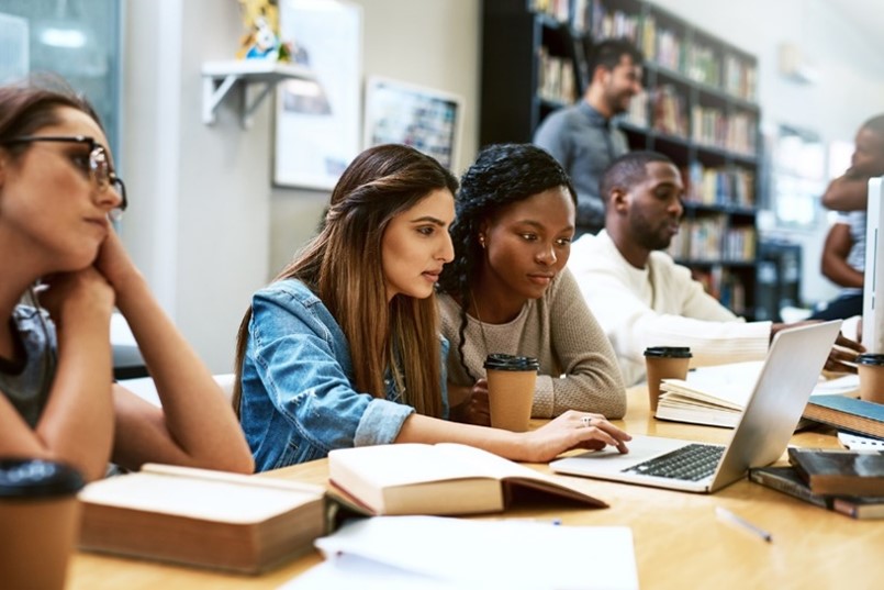 Four students sat studying at a long table. The two in the middle are working together on a laptop. Two other people stood at the back talking next to a bookcase