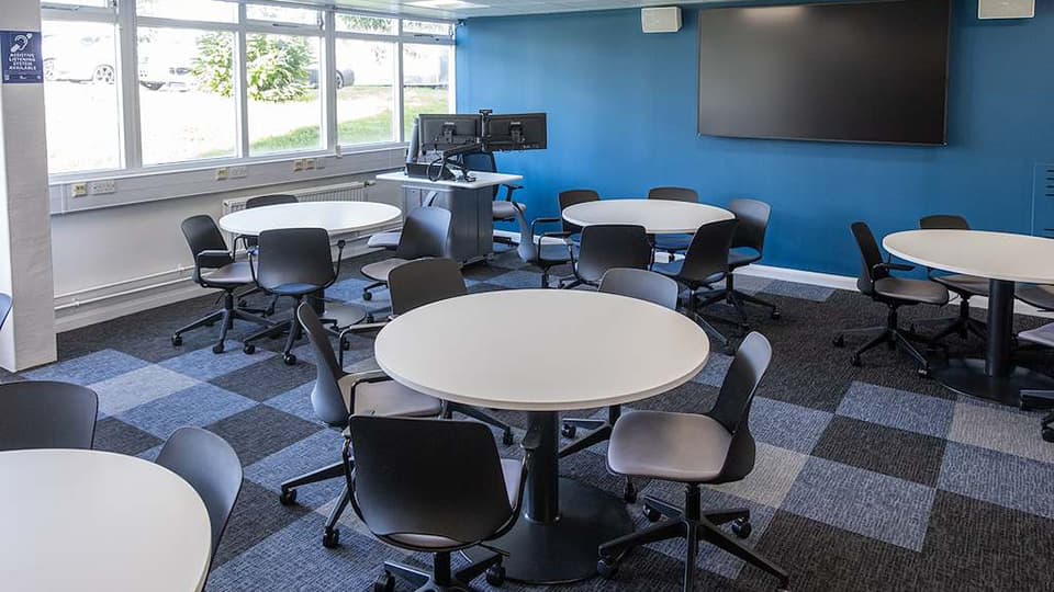 James France D003a - Flat teaching space on ground floor with natural light through big windows, blue walls, circular white tables and grey tiled carpet