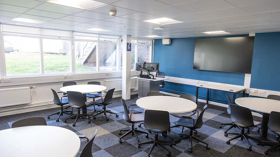 James France D003b - Flat teaching space on ground floor with natural light through big windows, blue walls, circular white tables and grey tiled carpet