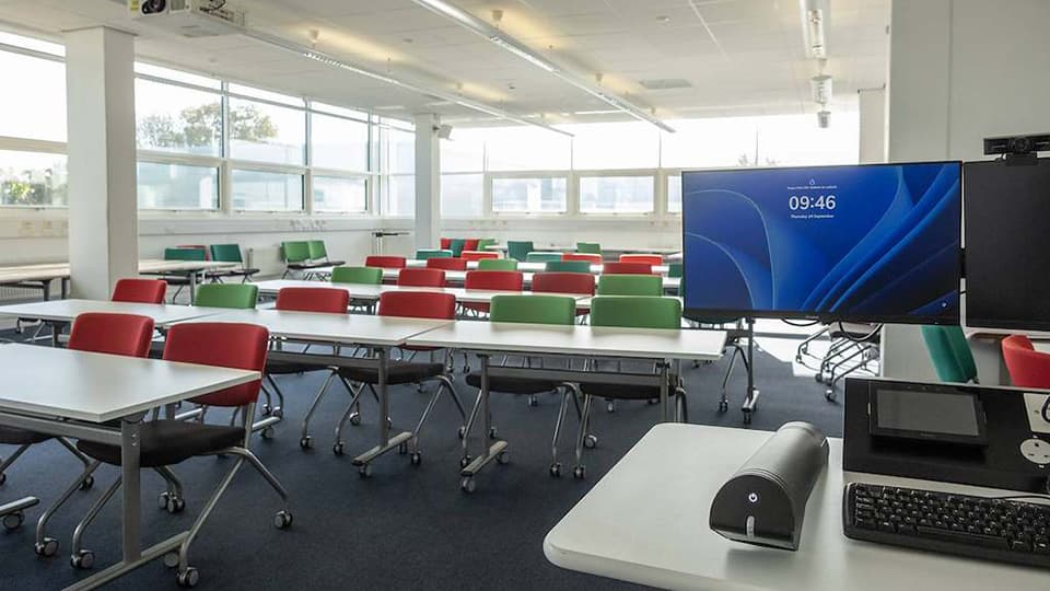 Brockington B114 - flat teaching space with white moveable desks, coloured chairs and windows on two walls.