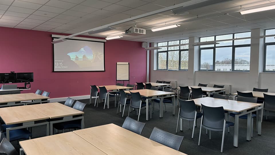A view of SCH138 in Schofield Building, a flat space with windows on both sides, pale desks and a mauve wall.