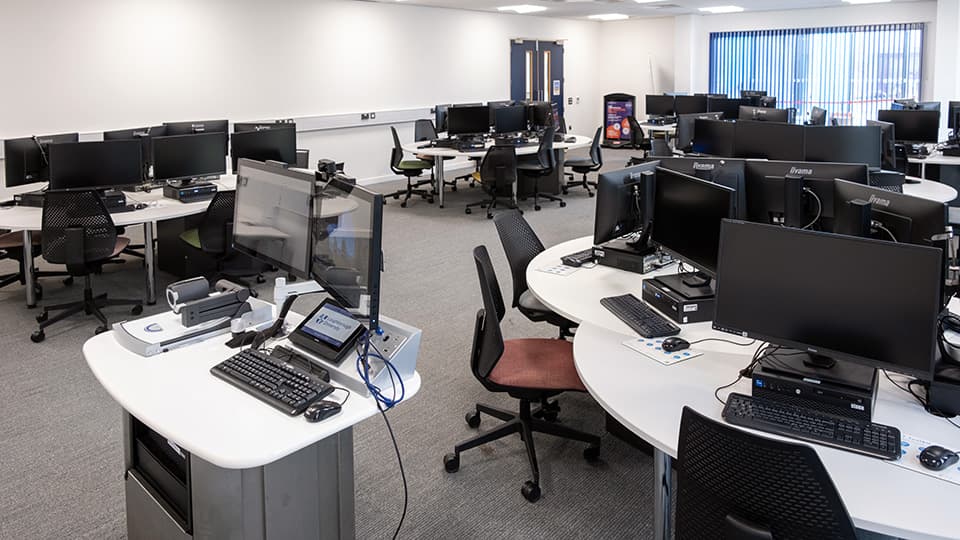 Computer teaching labs in West Park Labs - several white tables with groups of 6+ PCs in a white-walled space with grey carpet.