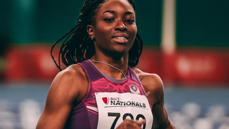 A close-up shot of a female track athlete with long braids, smiling while competing in a purple running kit with a 