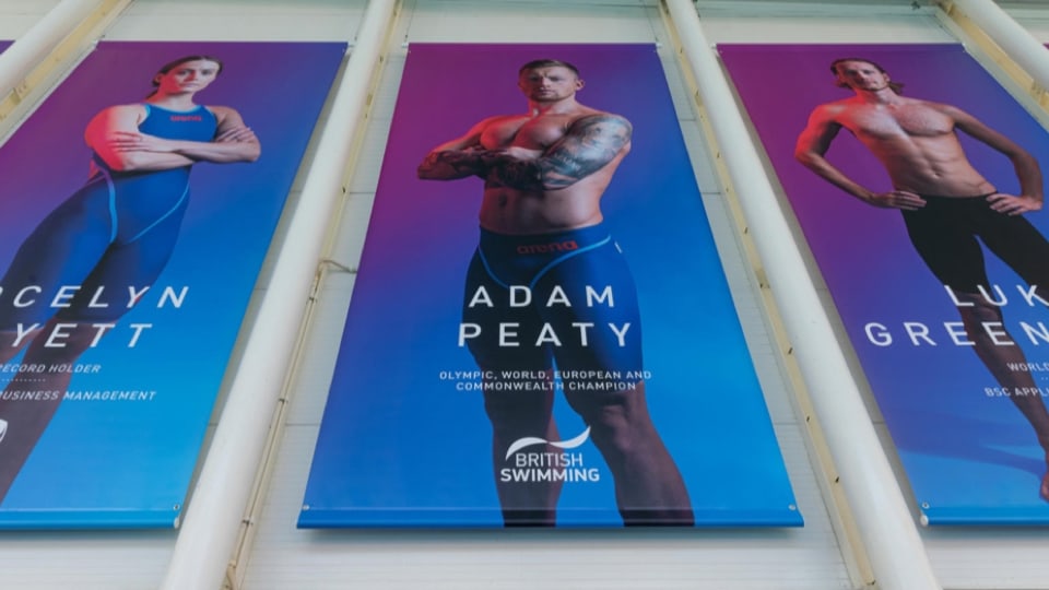 A low-angle view of three large promotional banners hanging side by side, featuring British swimmers in swimwear, including Olympic champion Adam Peaty in the centre.
