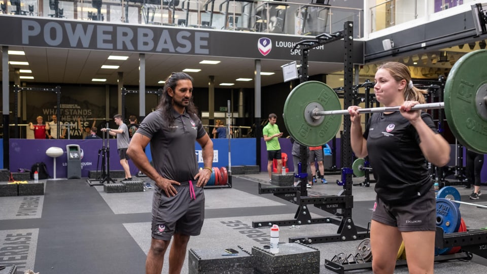 A female athlete lifts a barbell at a large, well-equipped indoor gym facility named 